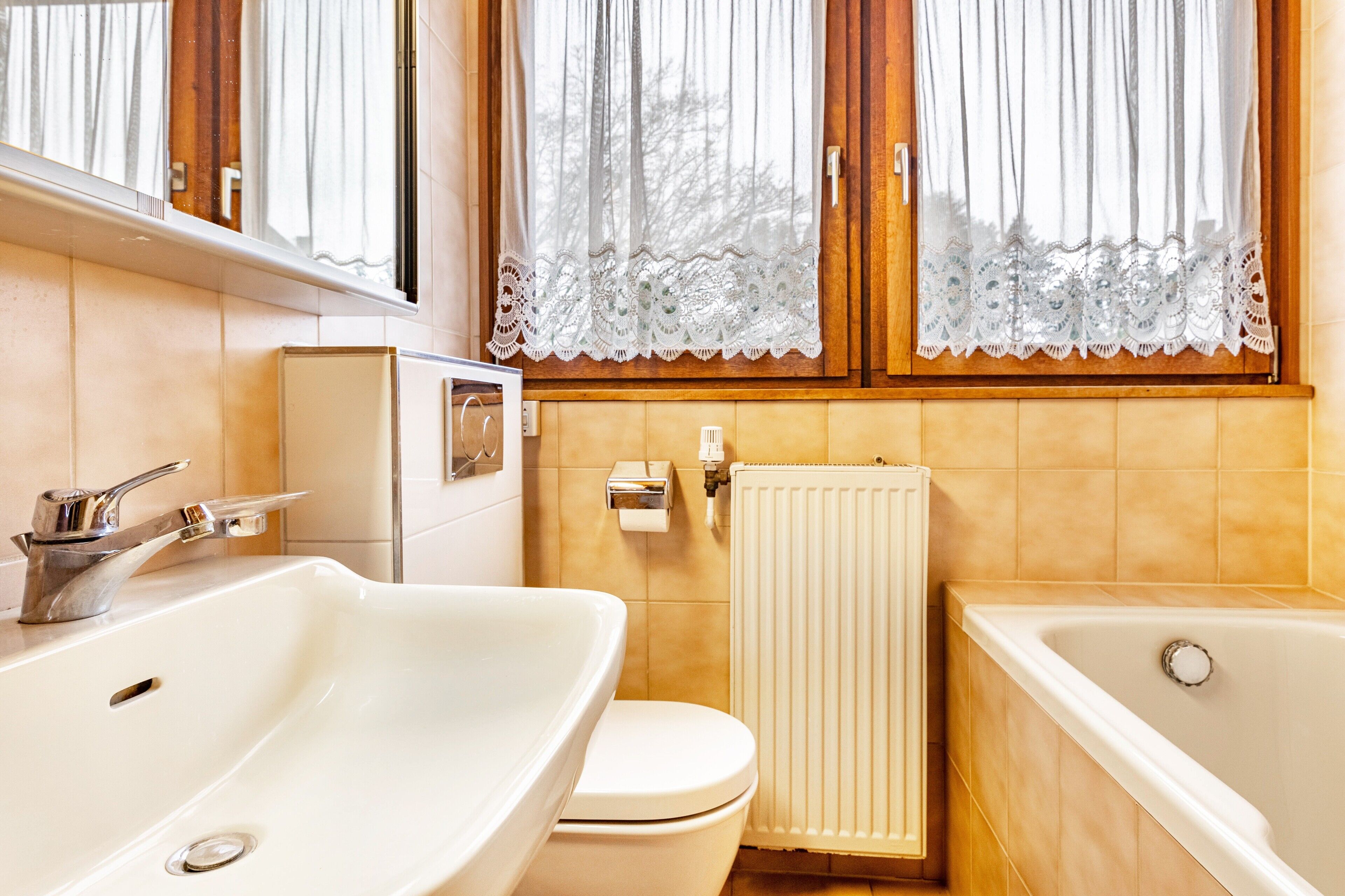 Ein Badezimmer mit Waschbecken, Toilette und Badewanne, einem Heizkörper und Spitzenvorhängen an den Fenstern.