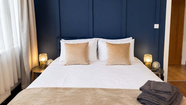 Une chambre d’hôtel avec un grand lit, deux tables de chevet avec des lampes, un plancher en bois et un mur bleu foncé orné d’un panneau décoratif original.