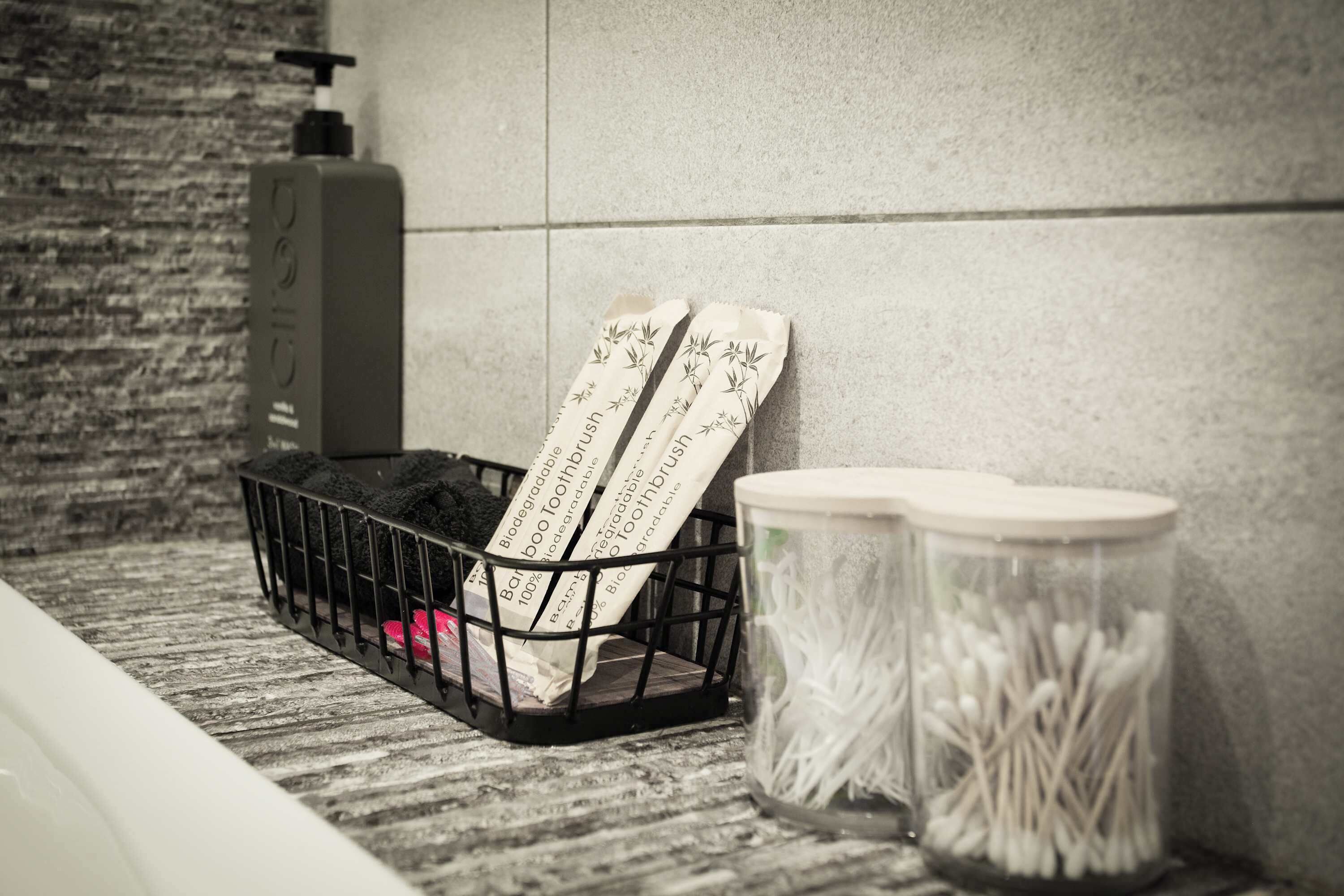 Una encimera de baño con una cesta negra que contiene cepillos de dientes y un recipiente transparente con bastoncillos de algodón.