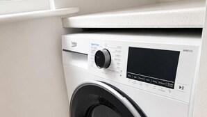 Un lave-linge à chargement frontal doté d’un panneau de commande, situé dans une pièce avec une fenêtre et un plancher en bois.