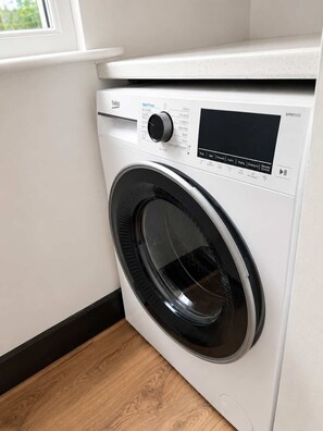 A front-loading washing machine with a control panel, located in a room with a window and wooden flooring.