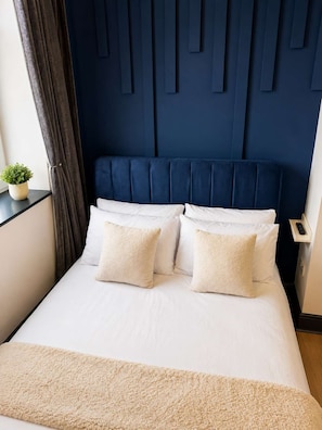 Cama com roupa de cama branca e almofadas bege, cabeceira azul escura e uma pequena planta em vaso sobre uma mesa de cabeceira.