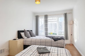 A bedroom with a bed, bedside table, lamp, and window with curtains. - Wycombe Oasis - Cozy 3-bed House Retreat (High Wycombe)