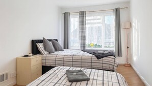 A bedroom with a bed, bedside table, lamp, and window with curtains. - Wycombe Oasis - Cozy 3-bed House Retreat (High Wycombe)