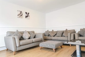 A living room with a grey sofa set, patterned cushions, a glass coffee table, and a wooden floor. - Wycombe Oasis - Cozy 3-bed House Retreat (High Wycombe)