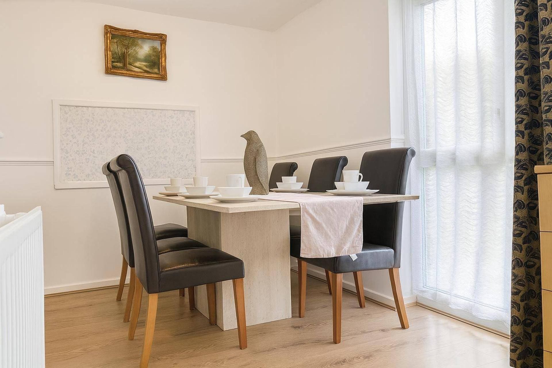 Ein Essbereich mit einem Tisch, gedeckt für vier Personen, je zwei Stühle an jeder Seite, einem gerahmten Bild an der Wand und einem Fenster mit Vorhängen.