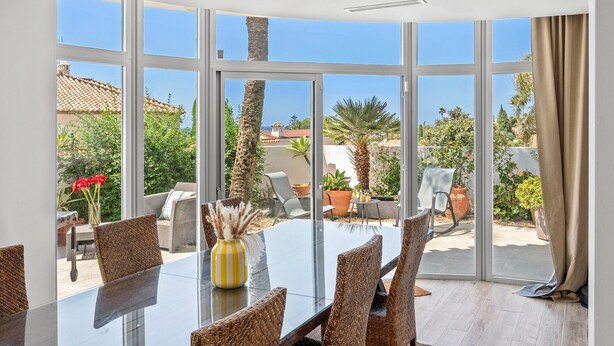 Un comedor con mesa de vidrio y sillas de mimbre, ventanas amplias con vistas a zonas verdes y edificios.