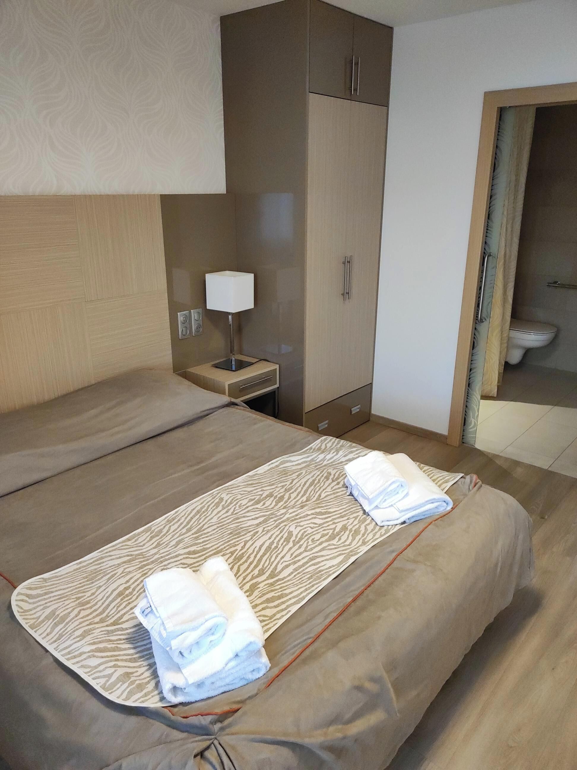 A hotel room with a bed, a nightstand, a lamp, a wardrobe, and a bathroom visible through an open door.