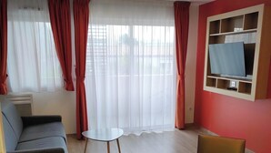 A modern living room with a sofa, a round table with chairs, a TV mounted on a wall, and a large window with sheer curtains. - Appart One Uzès (Uzès)
