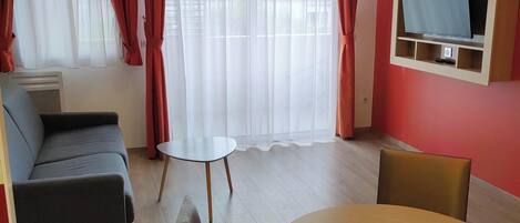 A modern living room with a sofa, a round table with chairs, a TV mounted on a wall, and a large window with sheer curtains.