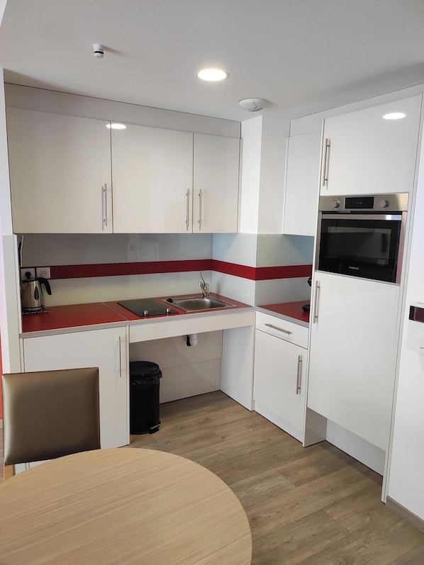 A compact kitchen with white cabinets, a red countertop, a built-in oven, and a sink. - Appart One Uzès (Uzès)