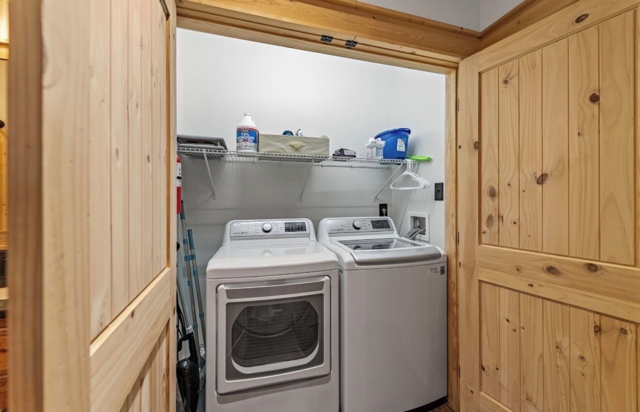 A laundry area with two washing machines, a shelf with cleaning supplies, and wooden cabinets.
