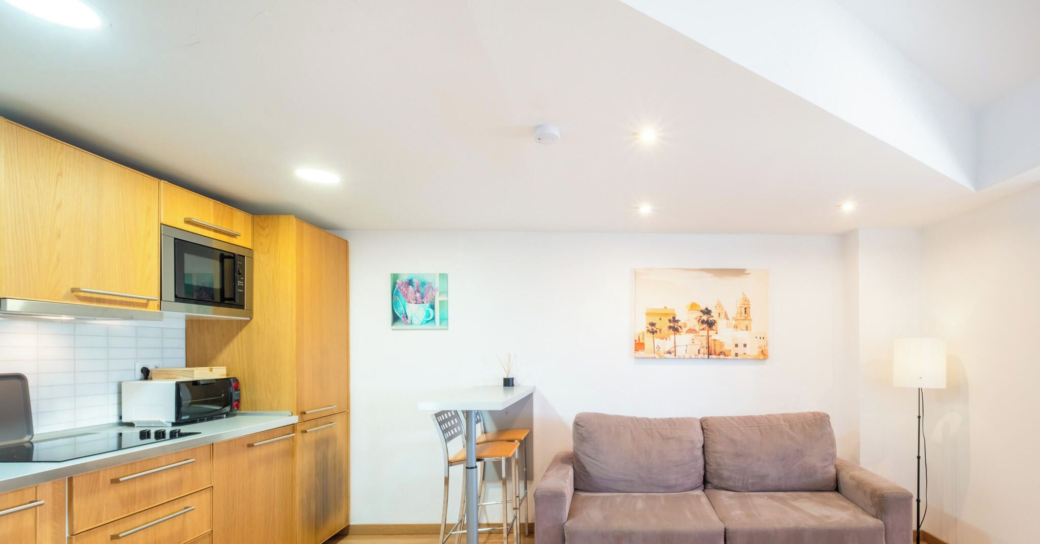 A compact kitchen with modern appliances, a dining area with a white table and metal chairs, and a sofa in a living space.
