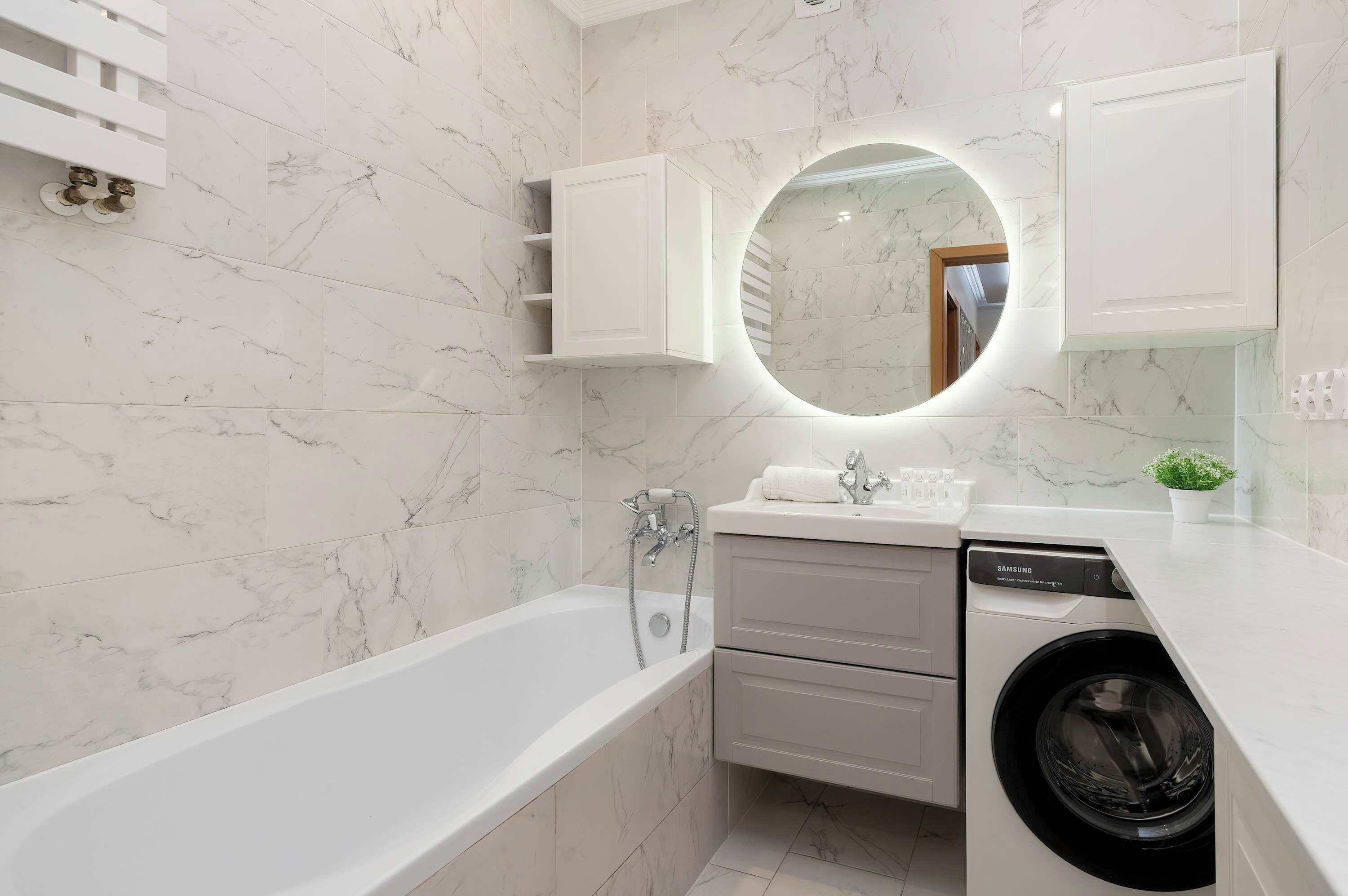 Een moderne badkamer met een witte marmeren wand, een ronde spiegel, een witte badkuip, een wasmachine en een wastafel met een wit aanrecht.