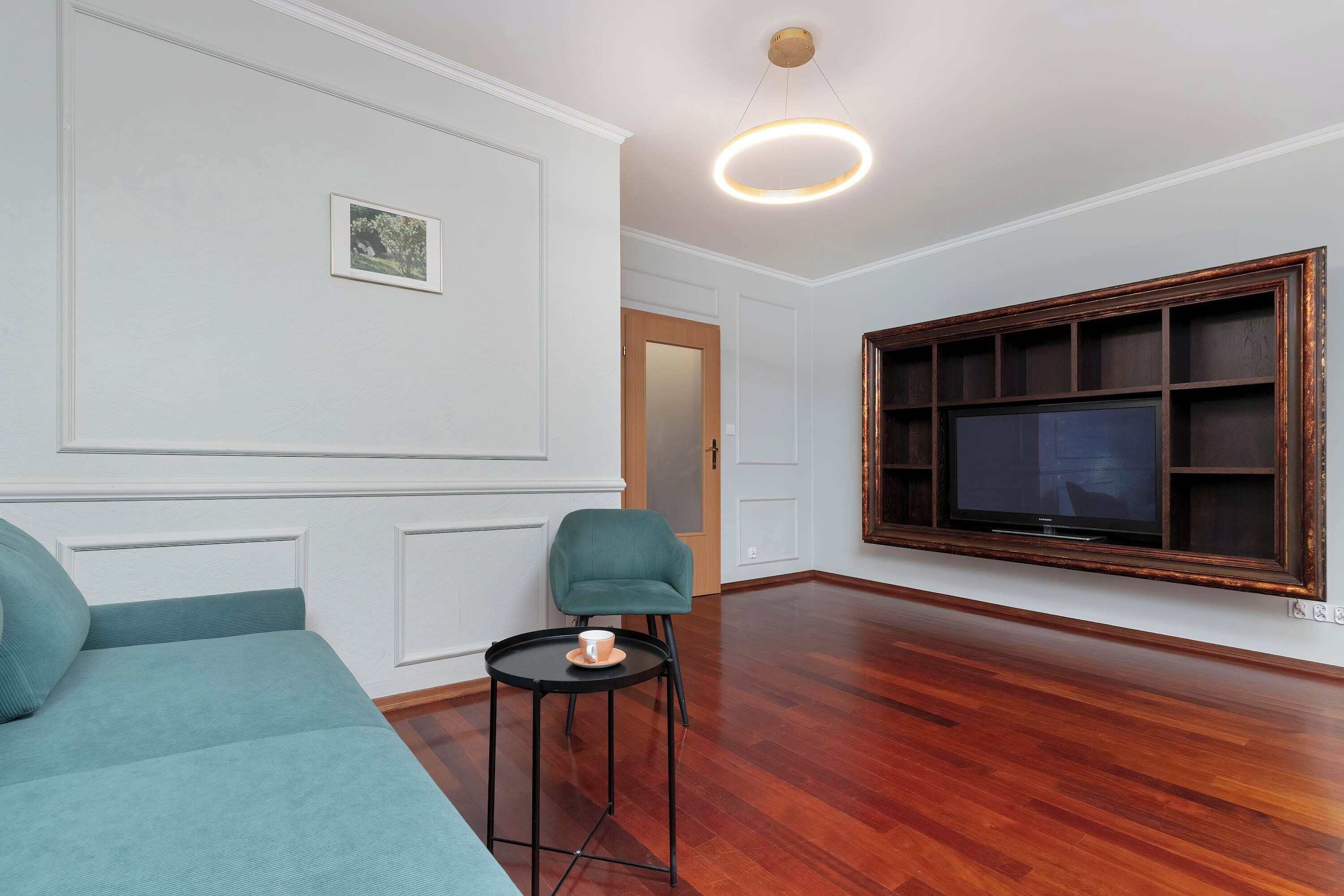 A room with a teal sofa, a small round table with a cup, a large dark-framed TV, and a wooden floor.