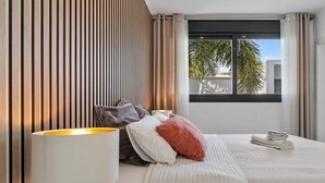 A hotel room with a bed, pillows, a lamp, and a window with a view of palm trees. - Modern Villa Lynx with Sauna, Heated Infinity Pool, and Garden (Las Lagunas de Mijas)