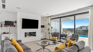 A modern living room with a fireplace, a large flat-screen TV, and a view of a pool and cityscape. - Modern Villa Lynx with Sauna, Heated Infinity Pool, and Garden (Las Lagunas de Mijas)