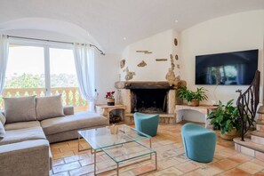 Ein Wohnzimmer mit einer Couch, zwei Sesseln, einem Glastisch, einem Kamin und einem an der Wand befestigten Fernseher.
