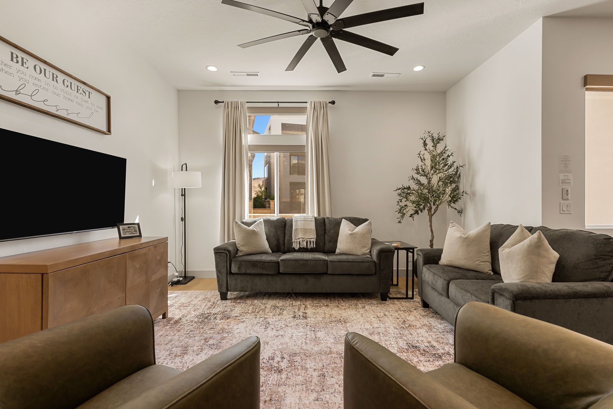 A modern living room with a flat-screen TV, two sofas, a coffee table, a floor lamp, and a ceiling fan.