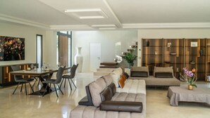 A modern living room with a sofa, dining table, and chairs. There is a large window, a painting, and a plant. - Villa spacieuse avec piscine (Opio)