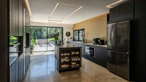 A modern kitchen with dark cabinetry, a central island, and a view to a patio area. - Villa spacieuse avec piscine (Opio)