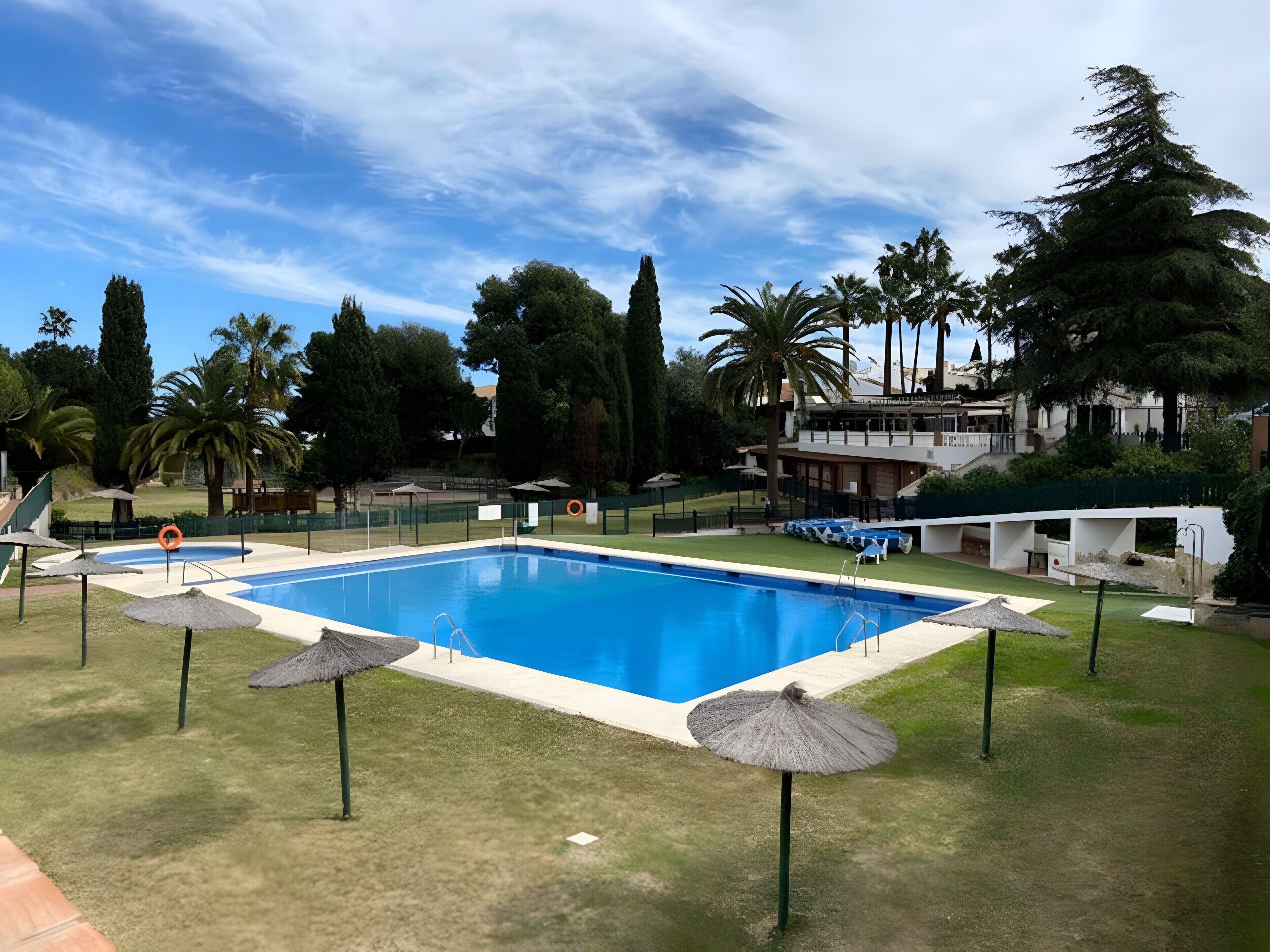 A swimming pool with a surrounding grassy area, umbrellas, and a building in the background.