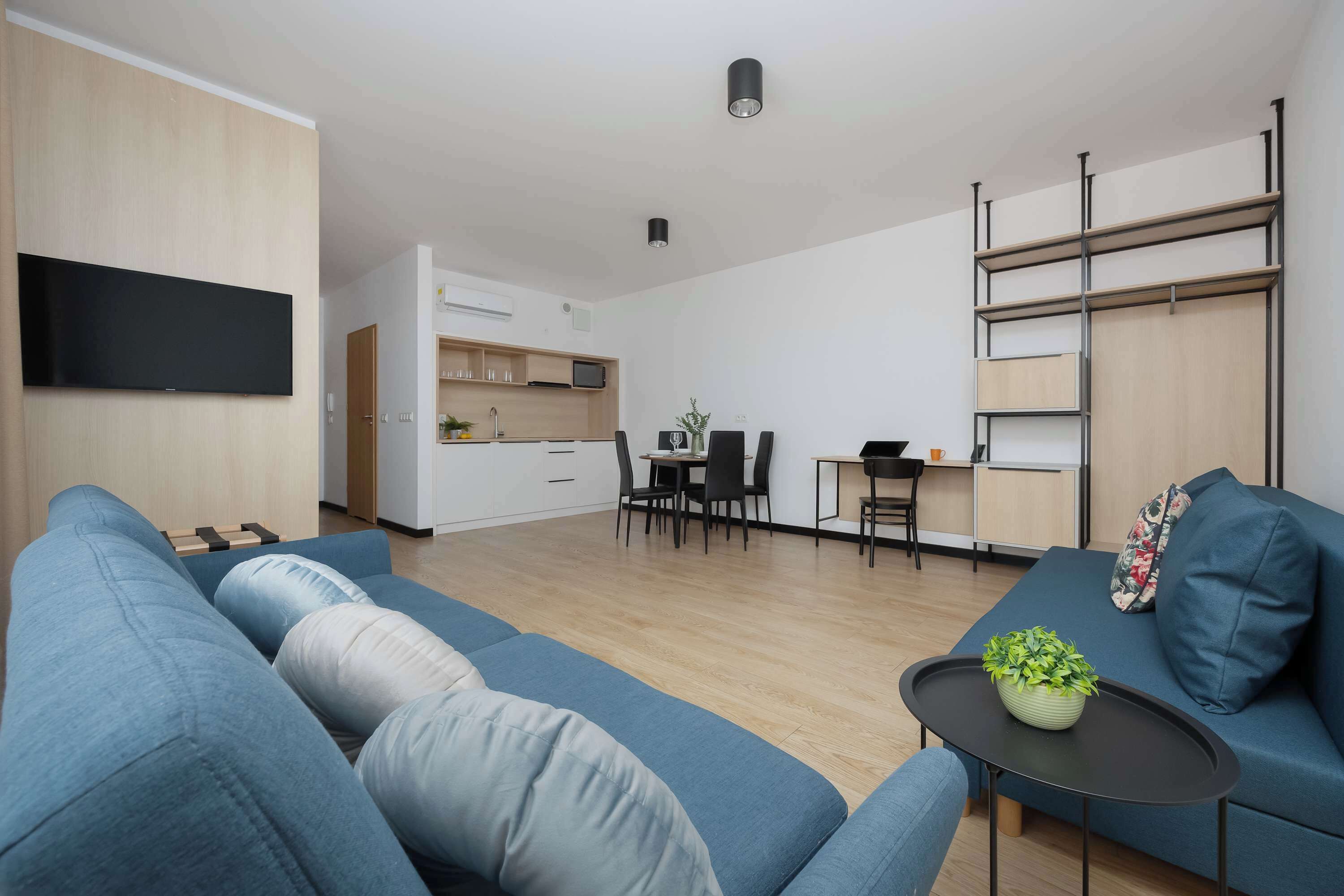 Un salon moderne avec un canapé bleu, une petite table sur laquelle est posée une plante, et un coin cuisine à l’arrière-plan.