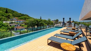 A rooftop pool with lounge chairs and umbrellas, surrounded by greenery and buildings. - Estúdio premium com varanda e sol da manhã SGR0217 (Florianópolis)