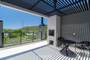 A balcony with a grill, a table, and chairs, overlooking a green landscape and a hill. - Estúdio premium com varanda e sol da manhã SGR0217 (Florianópolis)