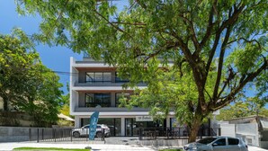 A modern two-story building with a visible restaurant sign, a car parked in front, and a large tree providing shade. - Estúdio premium com varanda e sol da manhã SGR0217 (Florianópolis)
