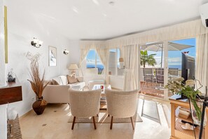 A spacious living room with a large window offering a view of the sea, a sofa, two armchairs, and a dining area with a table and chairs. - Spectacular Sea Views, Pool & Garden – Calahonda Apartment for 6 (Sitio de Calahonda)