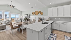 A modern kitchen with white cabinetry, a marble island, and a dining area with a table and chairs. - Saltwood Haven - Ocean Views w/ Elevator, Golf Cart, & Private Beach Access (Surf City)