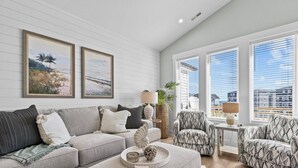 A modern living room with a grey sofa, patterned armchairs, and a coffee table with decorative items. There are two framed paintings on the wall and large windows with blinds. - Saltwood Haven - Ocean Views w/ Elevator, Golf Cart, & Private Beach Access (Surf City)