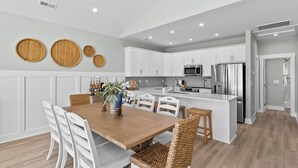 A modern kitchen with white cabinetry, a central island, and a dining area with a wooden table and white chairs. - Saltwood Haven - Ocean Views w/ Elevator, Golf Cart, & Private Beach Access (Surf City)