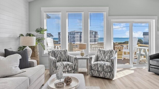 A modern living room with a large window, a sofa, two armchairs, a small table, and a view of the beach and buildings. - Saltwood Haven - Ocean Views w/ Elevator, Golf Cart, & Private Beach Access (Surf City)