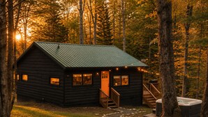 A cabin with a green roof, wooden exterior, and a porch with string lights. - Sauna, Hot Tub, Fire Pit & Creekside Deck | Niksen (Callicoon)