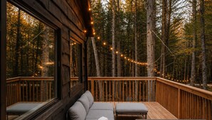 A wooden deck with a sofa and two chairs, surrounded by a forest with string lights. - Sauna, Hot Tub, Fire Pit & Creekside Deck | Niksen (Callicoon)