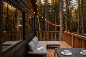 Eine Holzterrasse mit einer Couch und zwei Stühlen, umgeben von einem Wald mit Lichterketten.