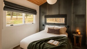 A bedroom with a bed, a window with a view of trees, a bedside table with a candle, and a hanging light fixture. - Sauna, Hot Tub, Fire Pit & Creekside Deck | Niksen (Callicoon)