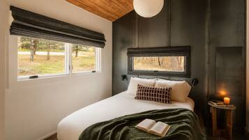 A bedroom with a bed, a window with a view of trees, a bedside table with a candle, and a hanging light fixture.