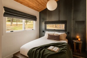 Ein Schlafzimmer mit Bett, Fenster mit Blick auf Bäume, Nachttisch mit Kerze und einer Pendelleuchte.