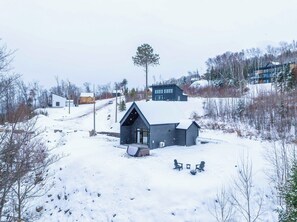 A snow-covered landscape with a modern house featuring a sloped roof and a small porch area with two chairs. - Modern Comfort | Spa & Mountain | Chalet ALBA (Saint-Côme)