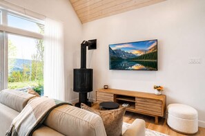 Ein Wohnzimmer mit Sofa, Kamin, Wandfernseher und einem Fenster mit Blick auf Grünflächen.