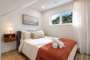 Ein Schlafzimmer mit Bett, Nachttisch, Lampe, einem Fenster mit Vorhängen und einem Bild an der Wand.