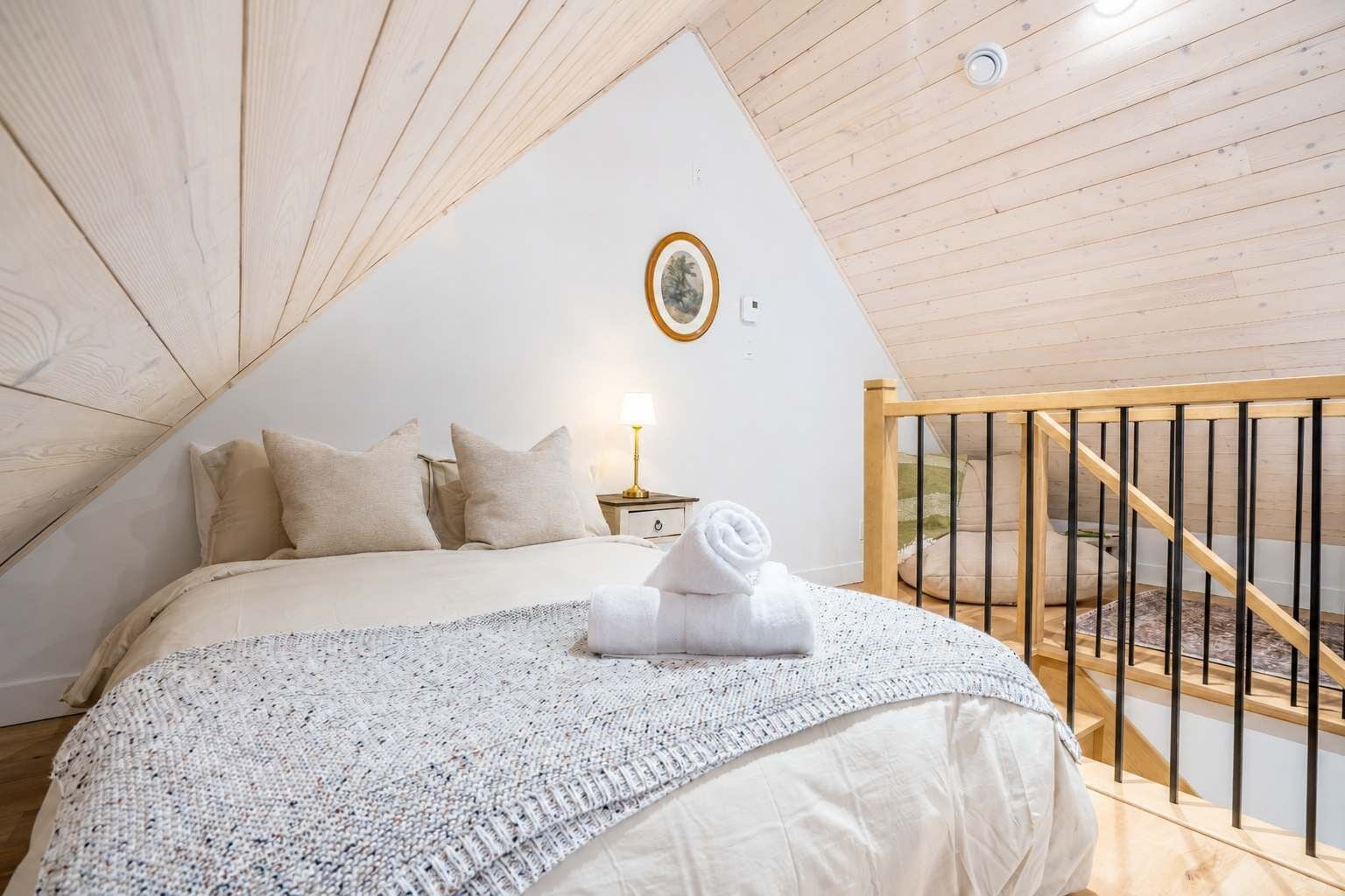 Een slaapkamer met een bed, kussens, een nachtkastje met een lamp, een lijst met een foto aan de muur, en een houten trap.