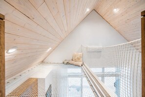 A sloped ceiling attic bedroom with a mesh safety railing, a cushioned bench, and a small window. - Modern Comfort | Spa & Mountain | Chalet ALBA (Saint-Côme)