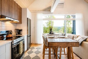 Eine moderne Küche mit Edelstahlgeräten, ein Essbereich mit Holztisch und -st ühlen sowie ein großes Fenster mit Blick ins Freie.