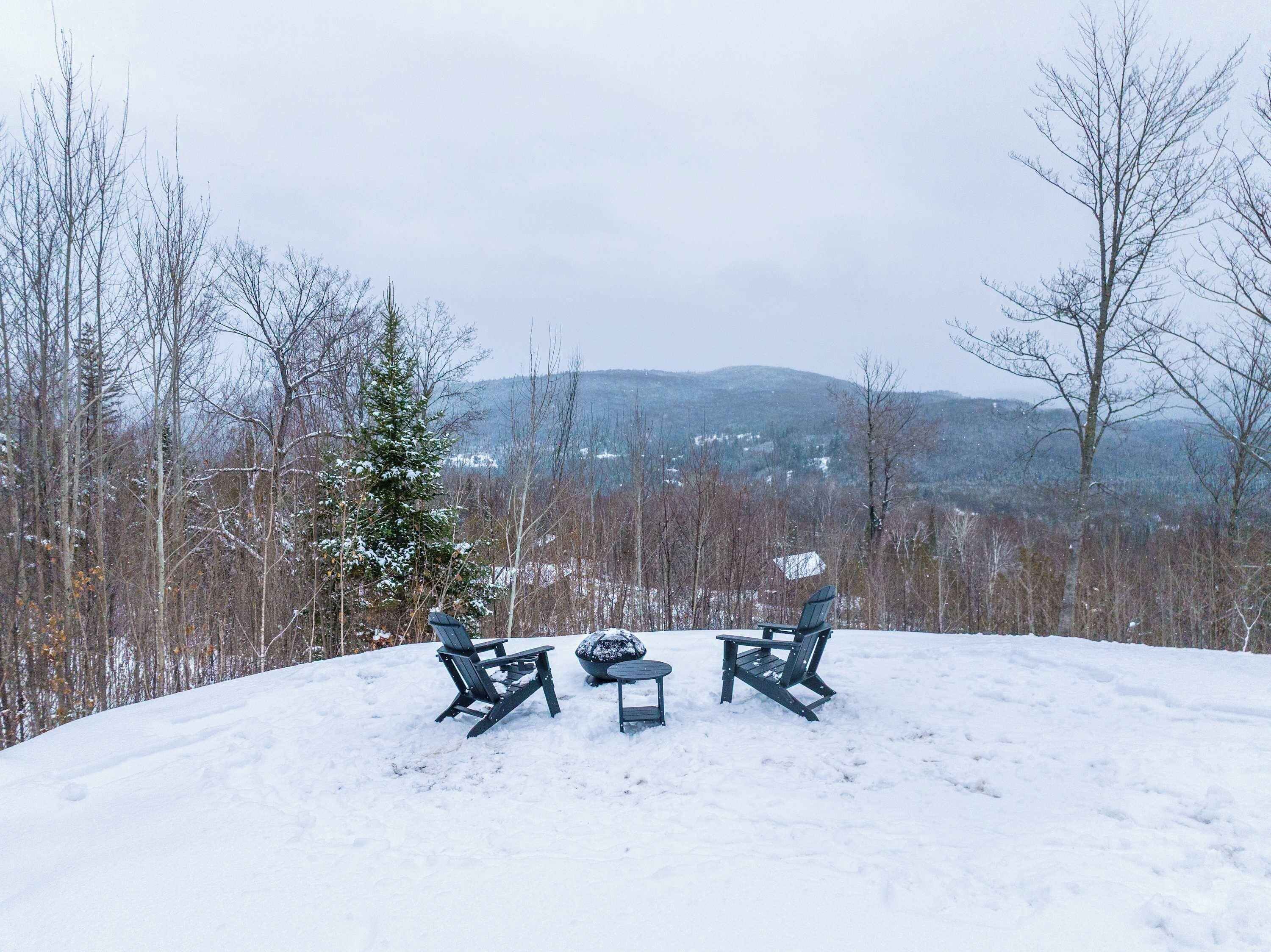 A snow-covered outdoor seating area with three Adirondack chairs and a small table, surrounded by trees and a view of distant hills.
