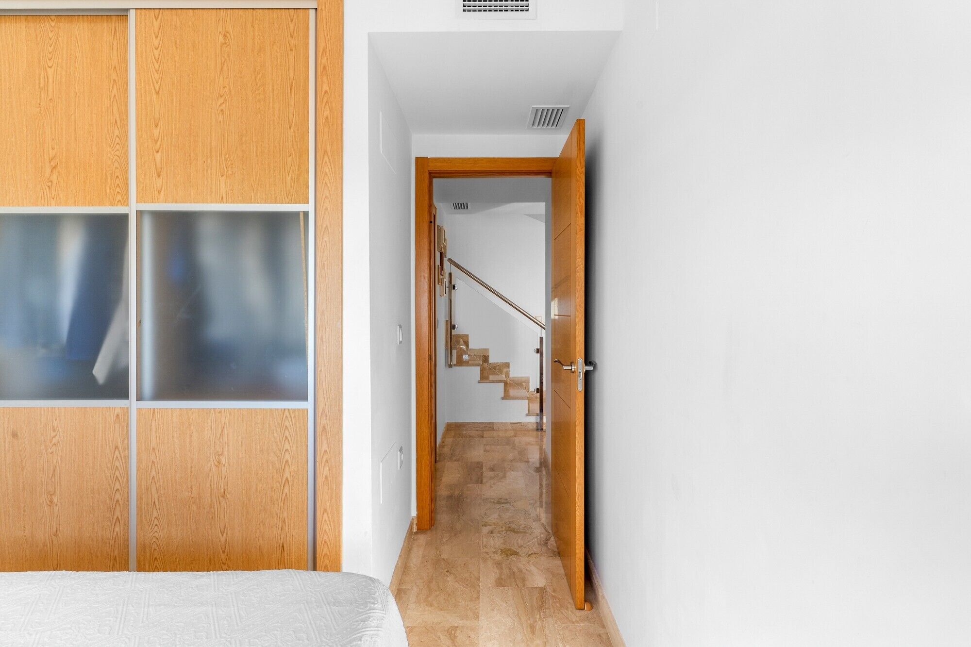 A narrow hallway with a wooden wardrobe, a bed, and a staircase leading to another room.