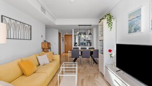 A modern living room with a sofa, a coffee table, a TV, and a dining area with chairs and a kitchen in the background.